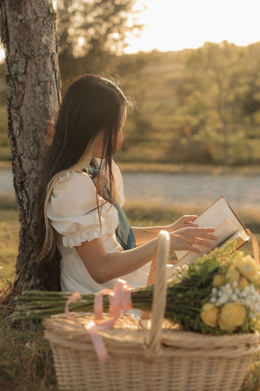 A romantic sunset picnic scene with fairy lights, a wicker basket, and yellow flowers beside a tree.
