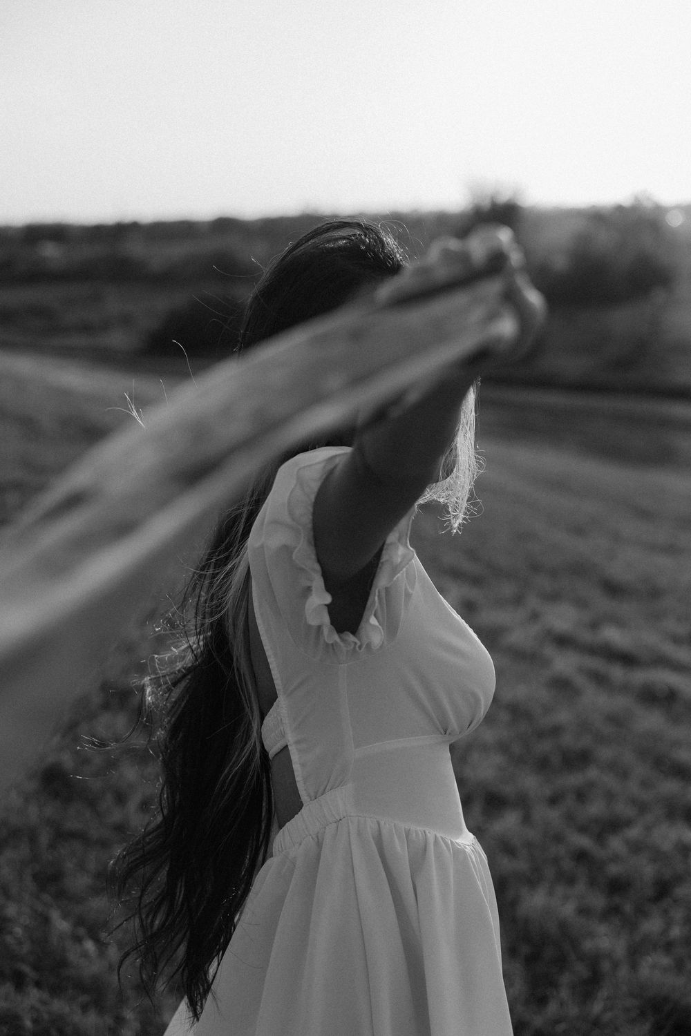 A person in a flowing white dress stands in a field at sunset with long dark hair blowing in the wind.