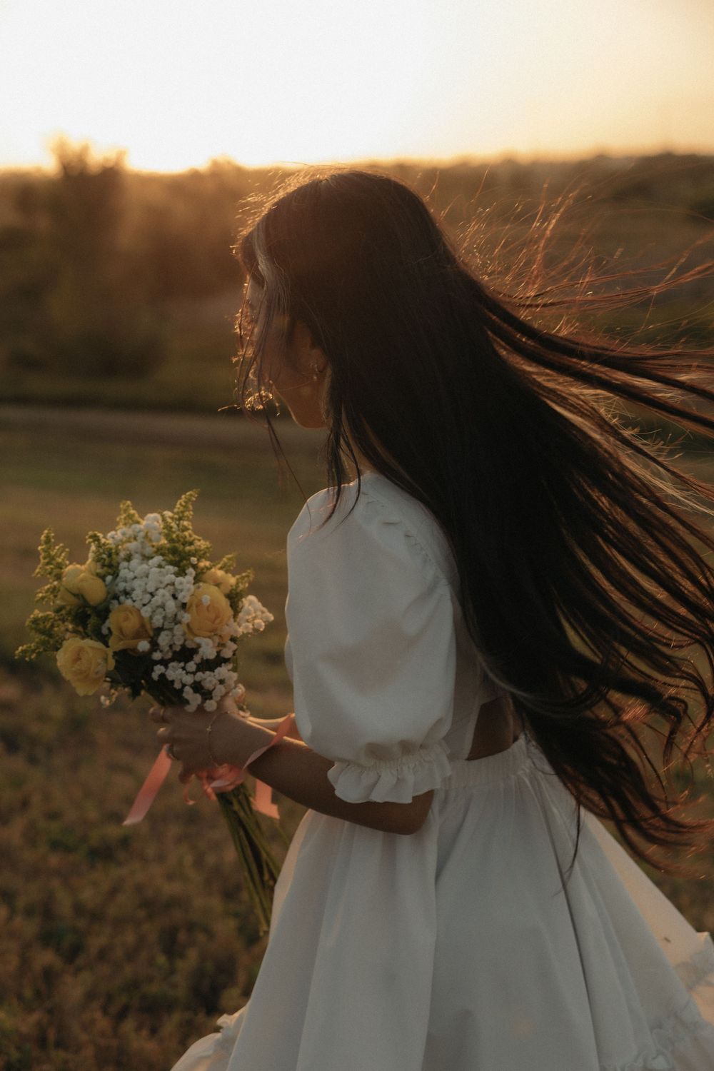 A figure in a flowing white dress holds a bouquet while their hair blows in the wind during golden hour sunset.