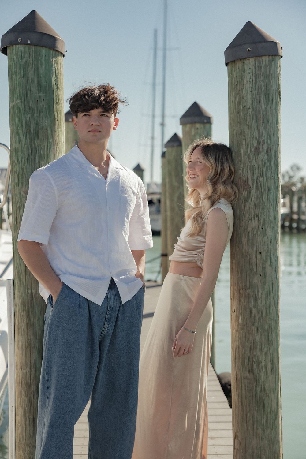 A couple stands together on a wooden dock by the water with wooden pilings in the background.