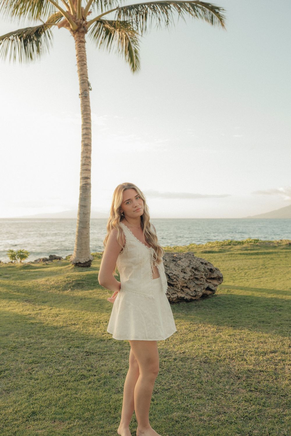 A woman in a white dress poses on a grassy lawn with palm trees and ocean views at sunset.