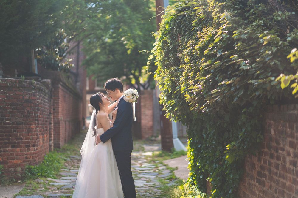 A newly married couple walking together through a lush garden pathway with brick walls and natural greenery.