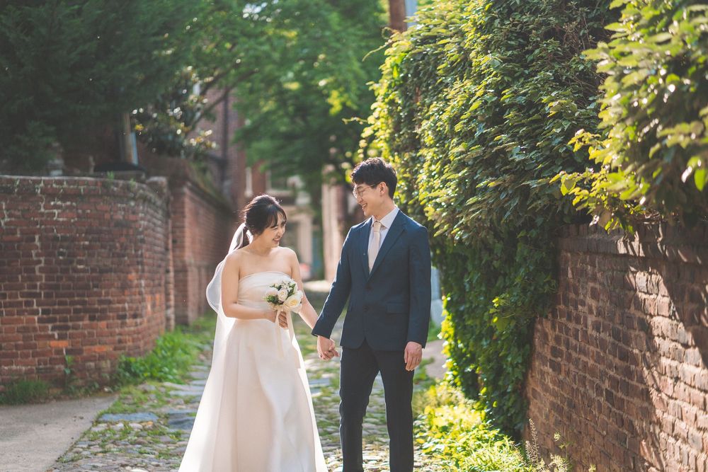 Couple in formal wedding attire poses together in a lush garden walkway with brick walls.