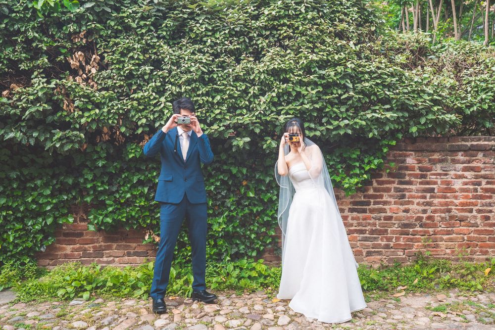 A couple in formal wedding attire poses in a garden against a brick wall with lush greenery in the background.