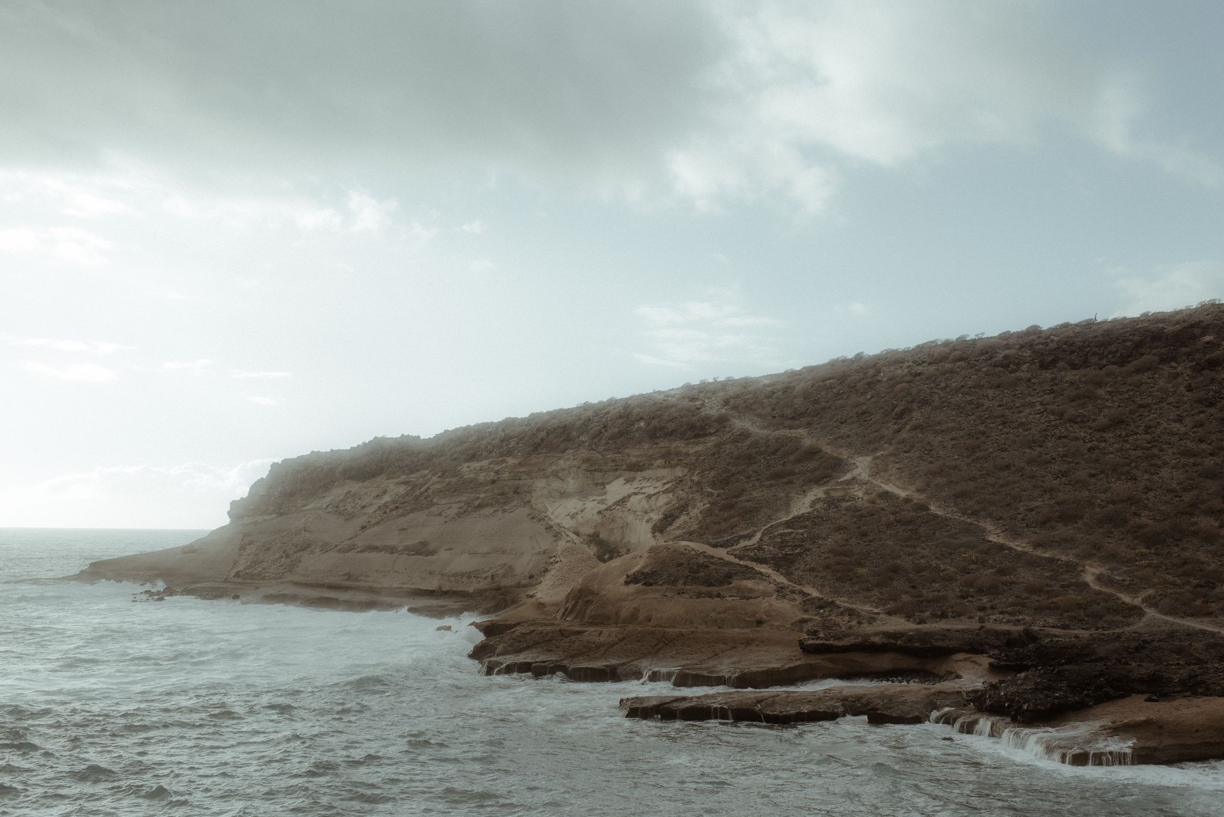 A rugged coastal cliff in La Caleta de Adeje with winding hiking trails overlooks a misty Atlantic Ocean under a moody, cloudy sky.
