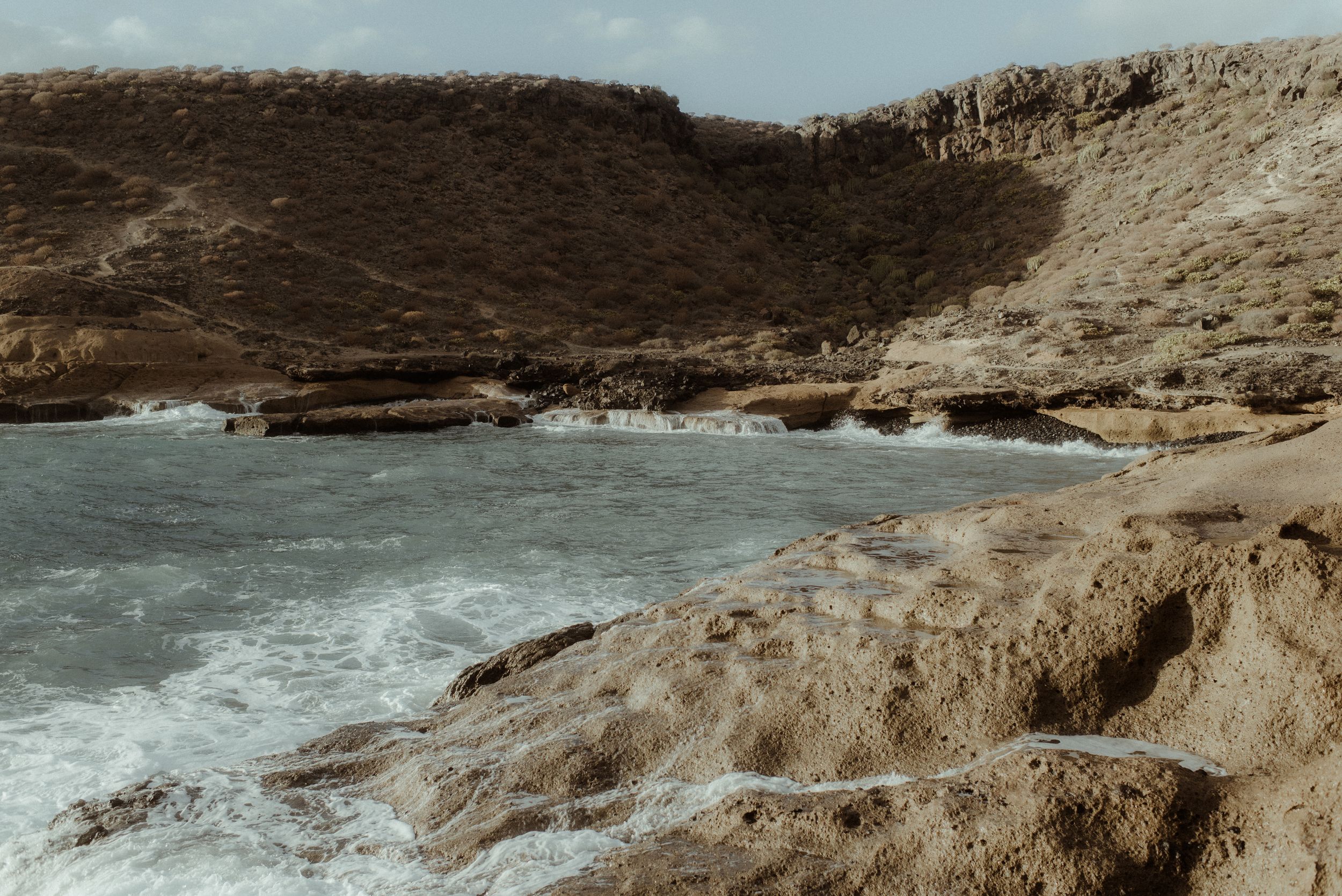 Rugged coastal cliffs of Playa de los Morteros meet ocean waves along a rocky desert shoreline under an overcast sky.