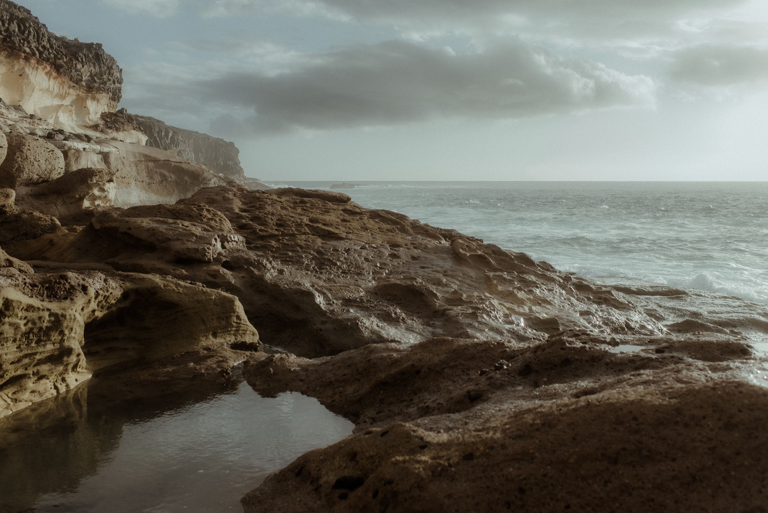 Dramatic coastal landscape with rugged cliffs and tidal pools against a moody ocean backdrop at sunset in La Caleta de Adeje, Tenerife.
