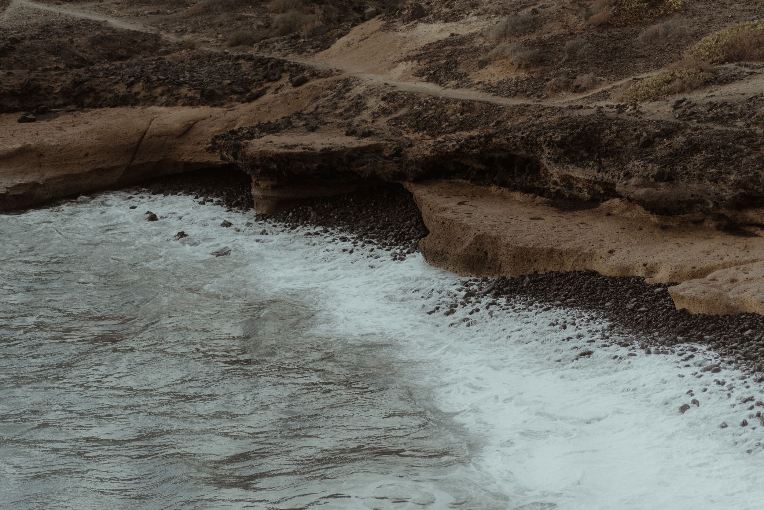 Rocky coastal shoreline of Playa de los Morteros with dark waves crashing against brown cliffs and scattered rocks along the water's edge.