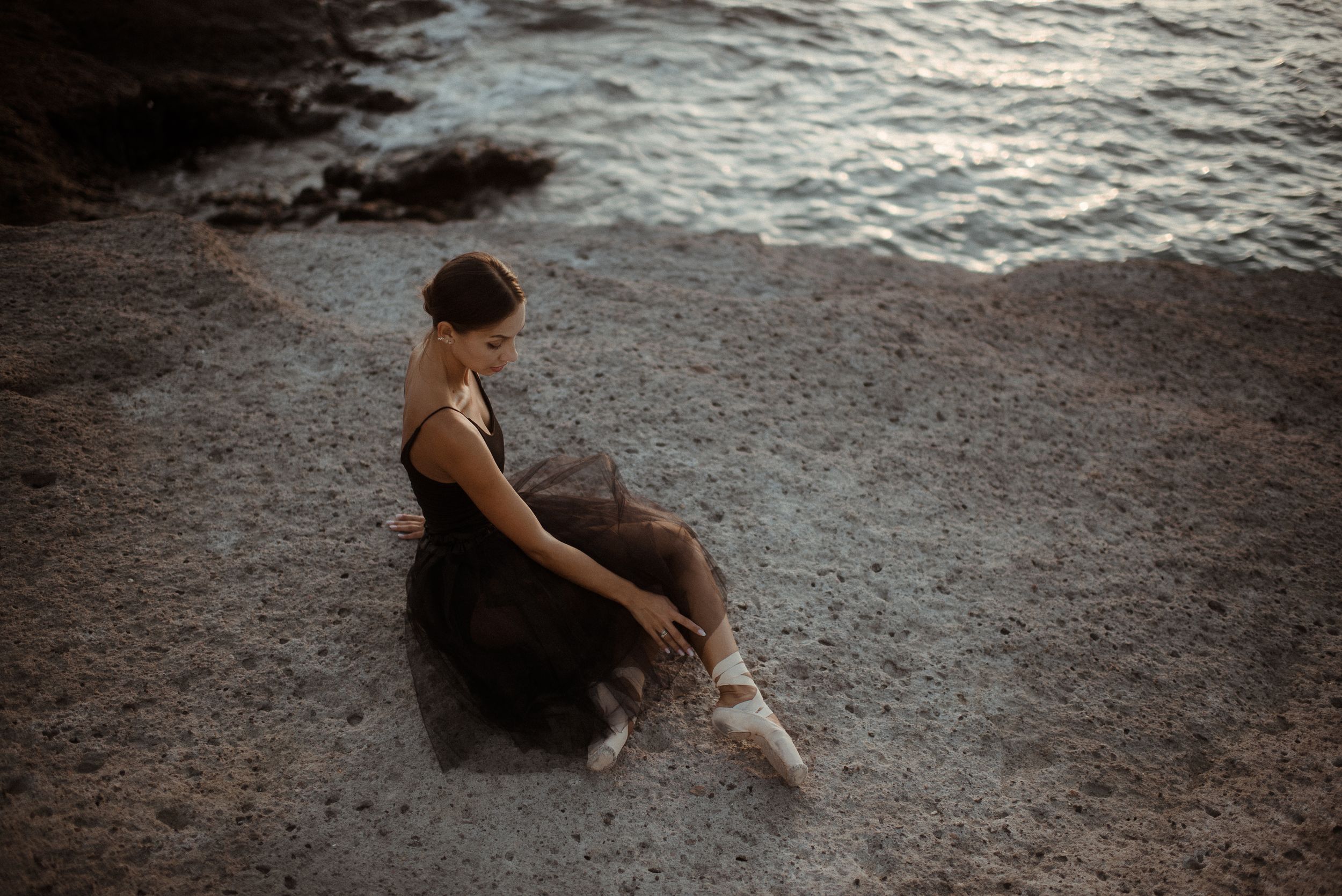 Ballerina in black leotard sits gracefully on rocky beach at sunset as waves lap against the shore in La Caleta de Adeje, Tenerife.