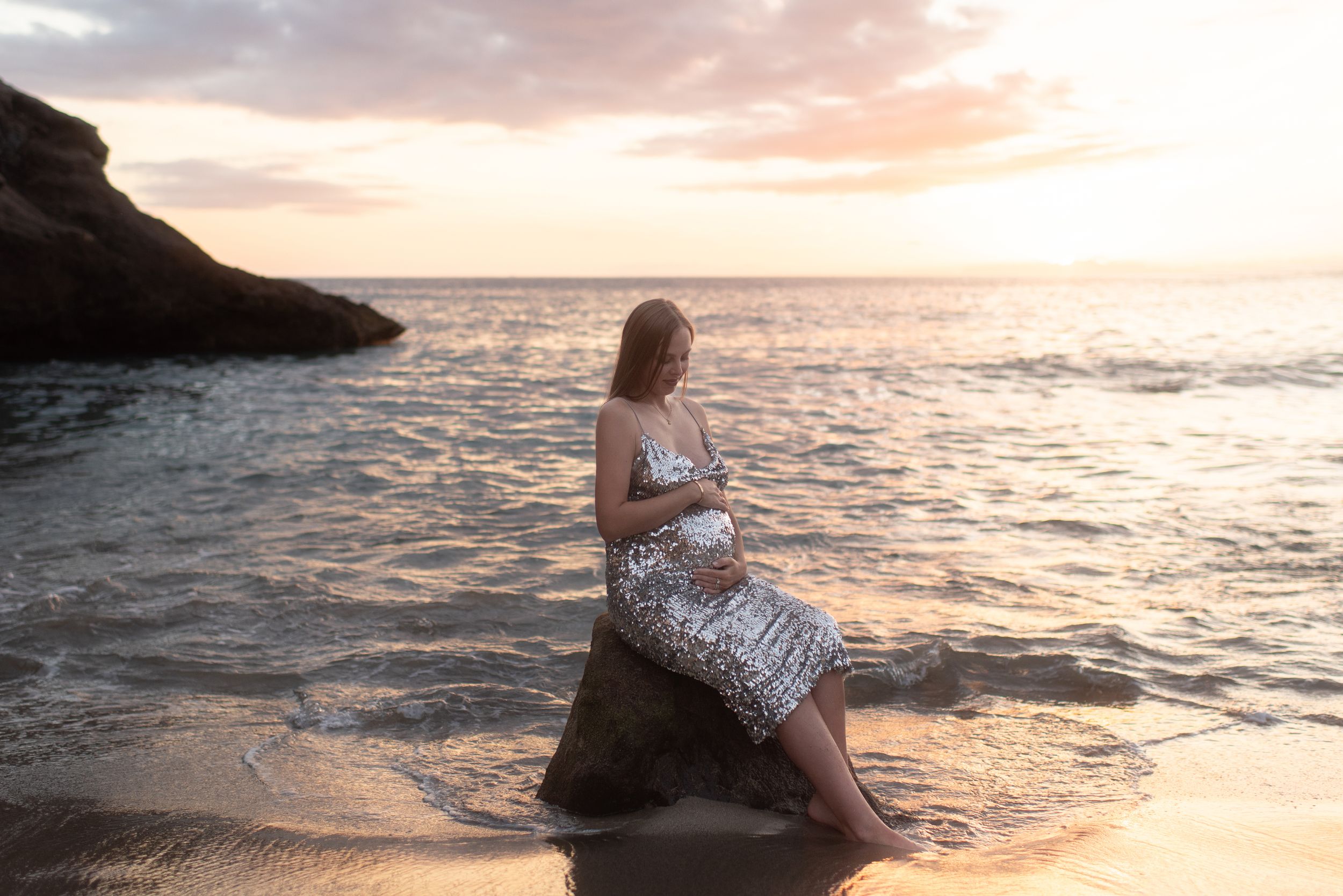 A pregnant beauty sitting on coastal rocks watching the sunset over the ocean horizon at Costa Adeje.