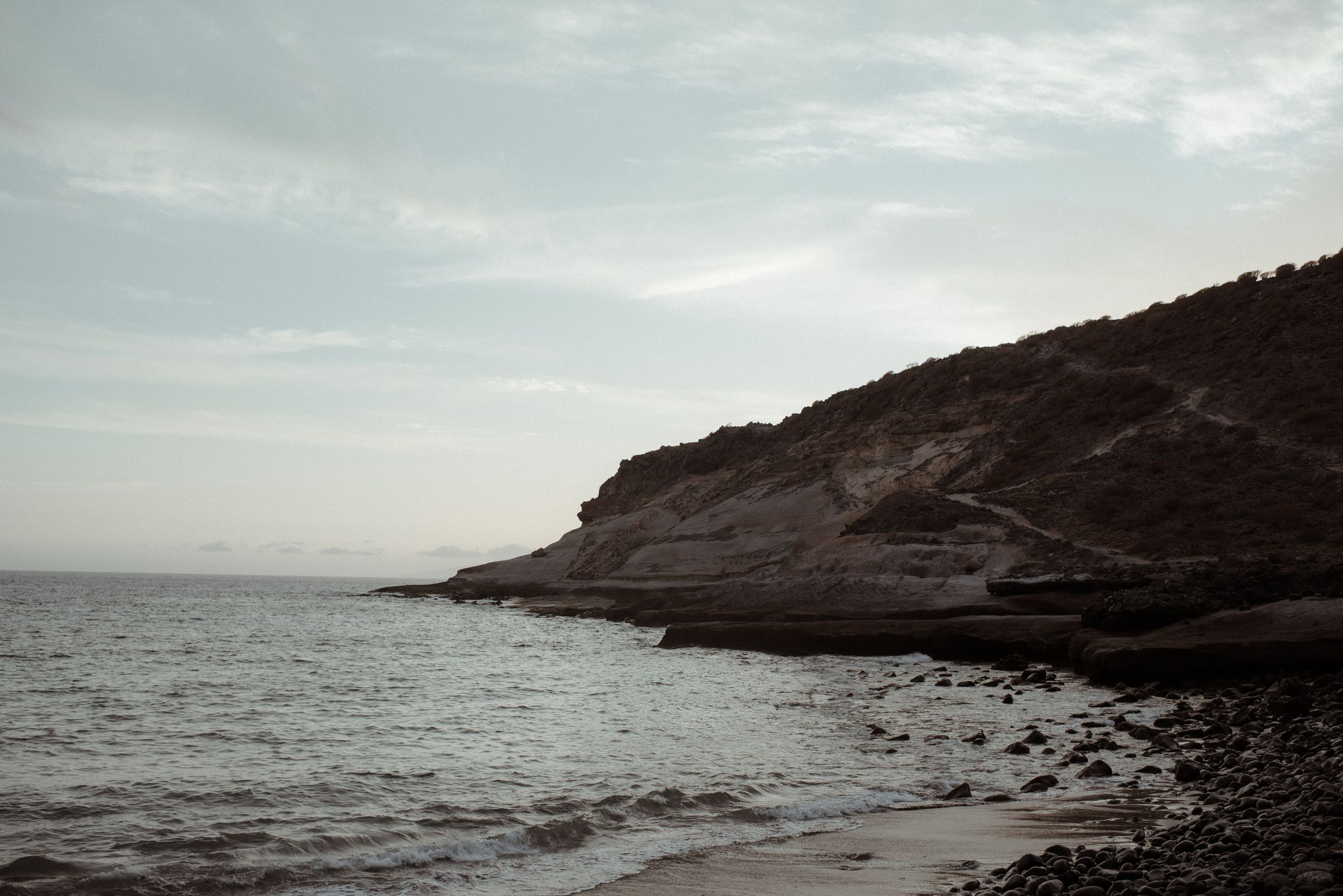 A rocky coastline with cliffs meets the sea under an overcast sky, creating a moody monochrome landscape in La Caleta de Adeje, Tenerife.