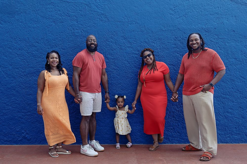 Group stands in line against vibrant blue wall wearing casual orange, red and white outfits at their family portraits in Cancun.