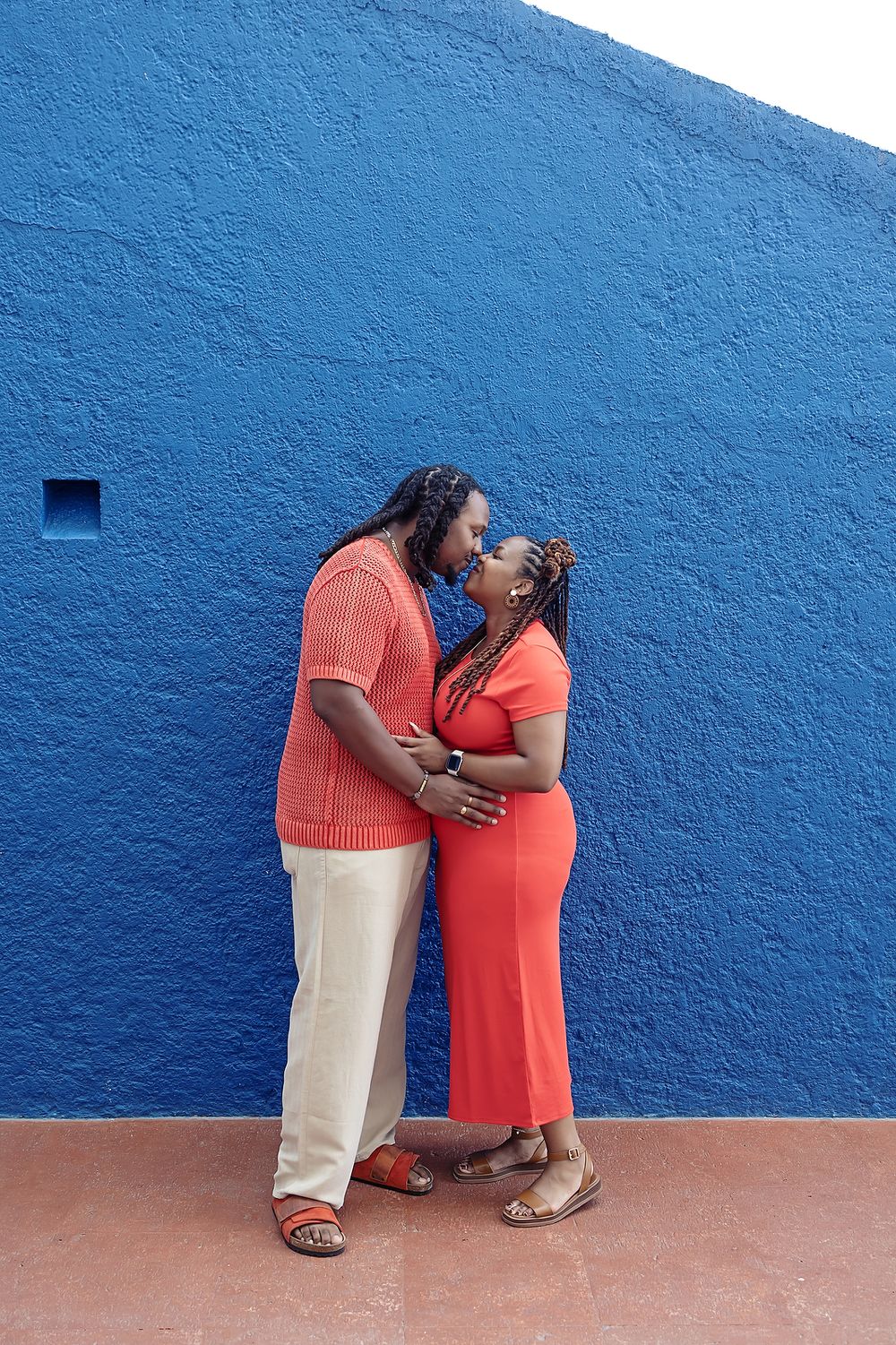 Two people wearing contrasting coral and beige outfits lean against a vibrant blue wall, creating a warm artistic composition.
