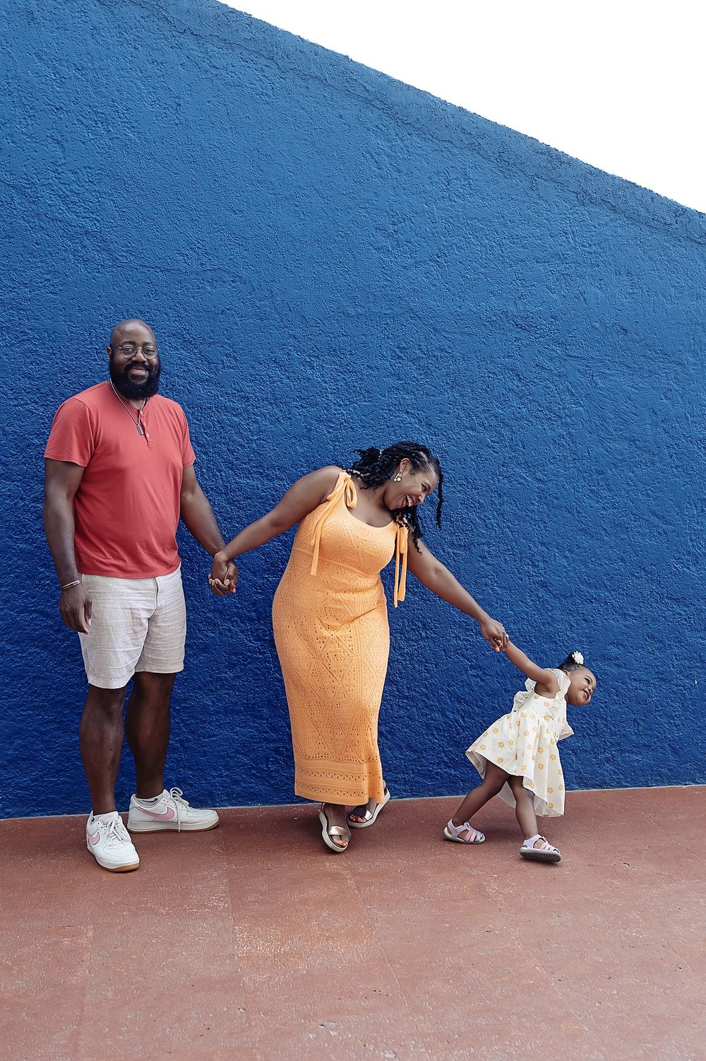 Tender sequence showing adults in coral and orange outfits interacting with a small child in white against a blue wall.
