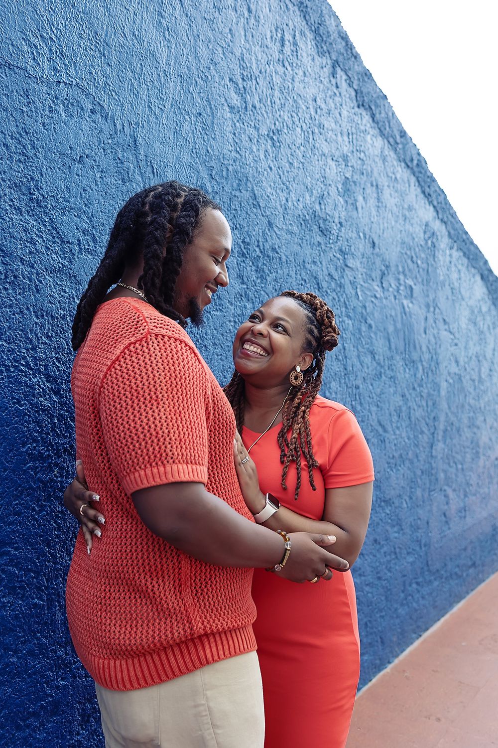 A couple in matching coral outfits share a loving embrace against a vibrant blue wall background.