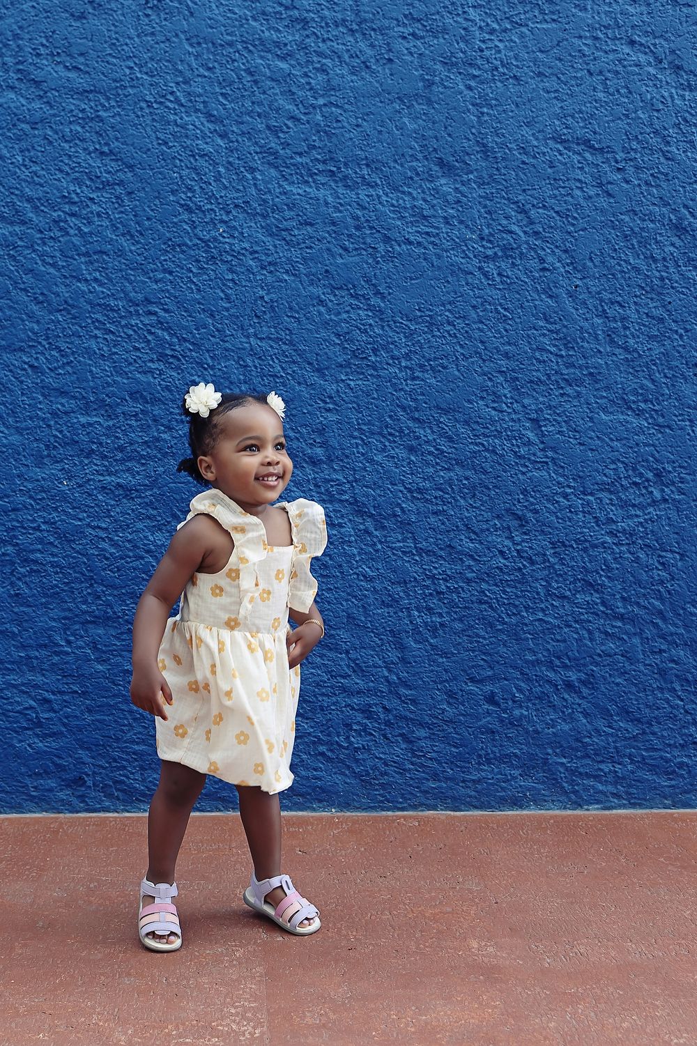 A young child wears a yellow floral dress and white sandals while posing happily against a bright blue wall.