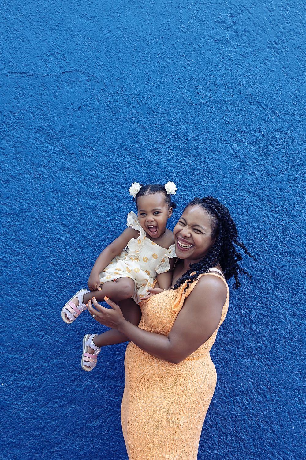 Person wearing orange dress poses joyfully with small child against vibrant blue wall background.