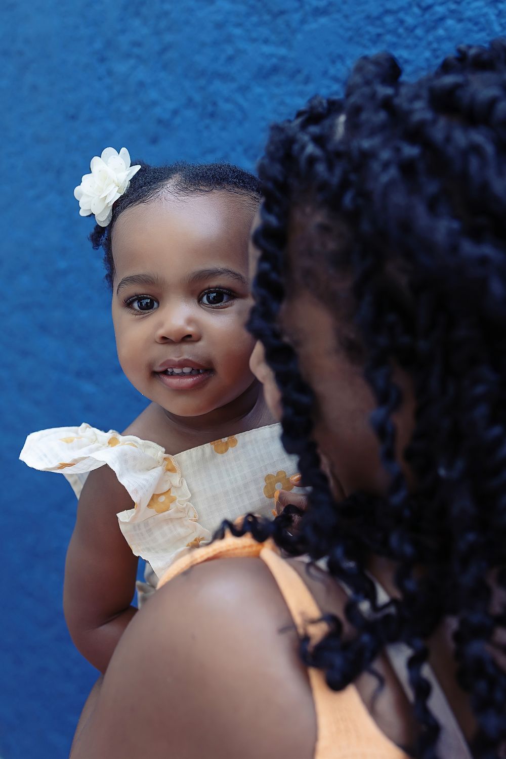 Child in white dress smiles while being held against vibrant blue wall, wearing white flower hair accessory.