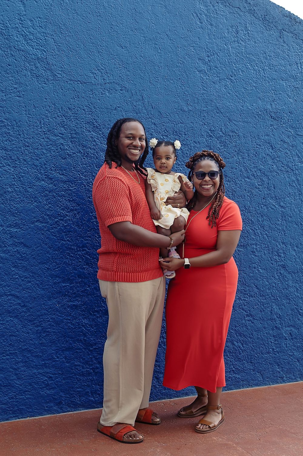 Against a vibrant blue wall, figures in red and beige outfits share warm embraces while seated on a brown tiled floor.