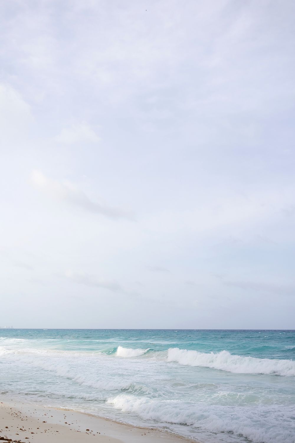 Tranquil ocean waves roll onto a sandy beach under an overcast sky with turquoise waters stretching to the horizon.