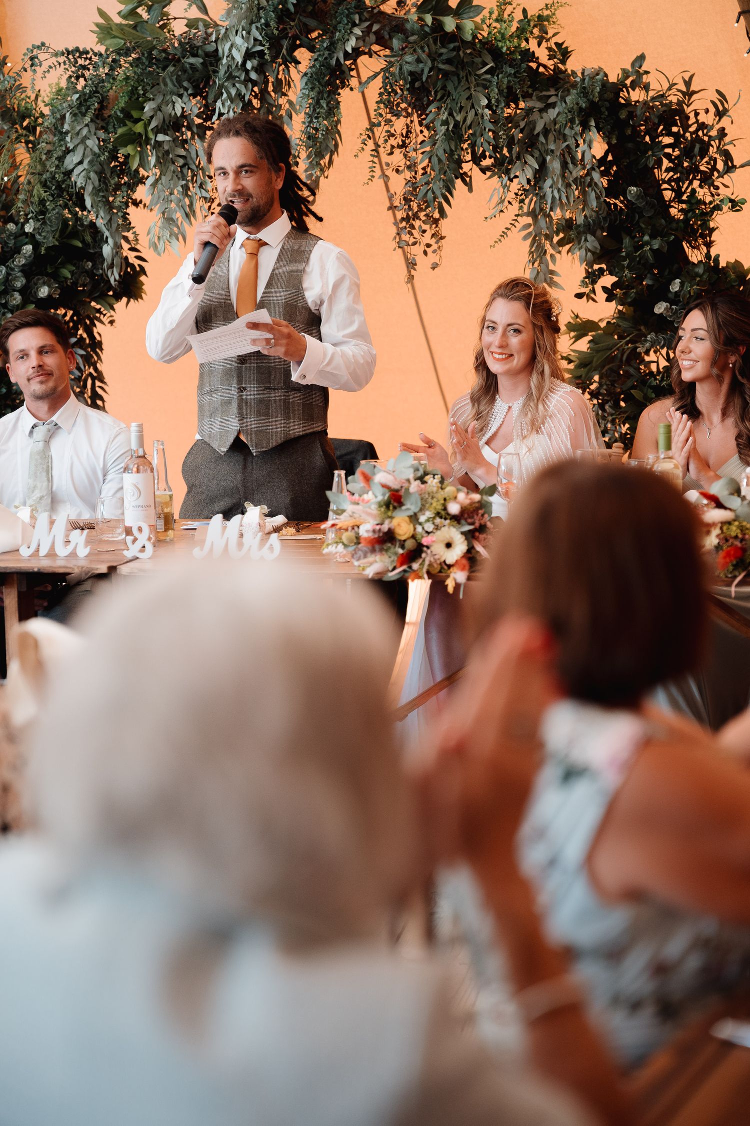 Wedding reception celebration under elegant greenery arch with table decor and floral arrangements during speeches.