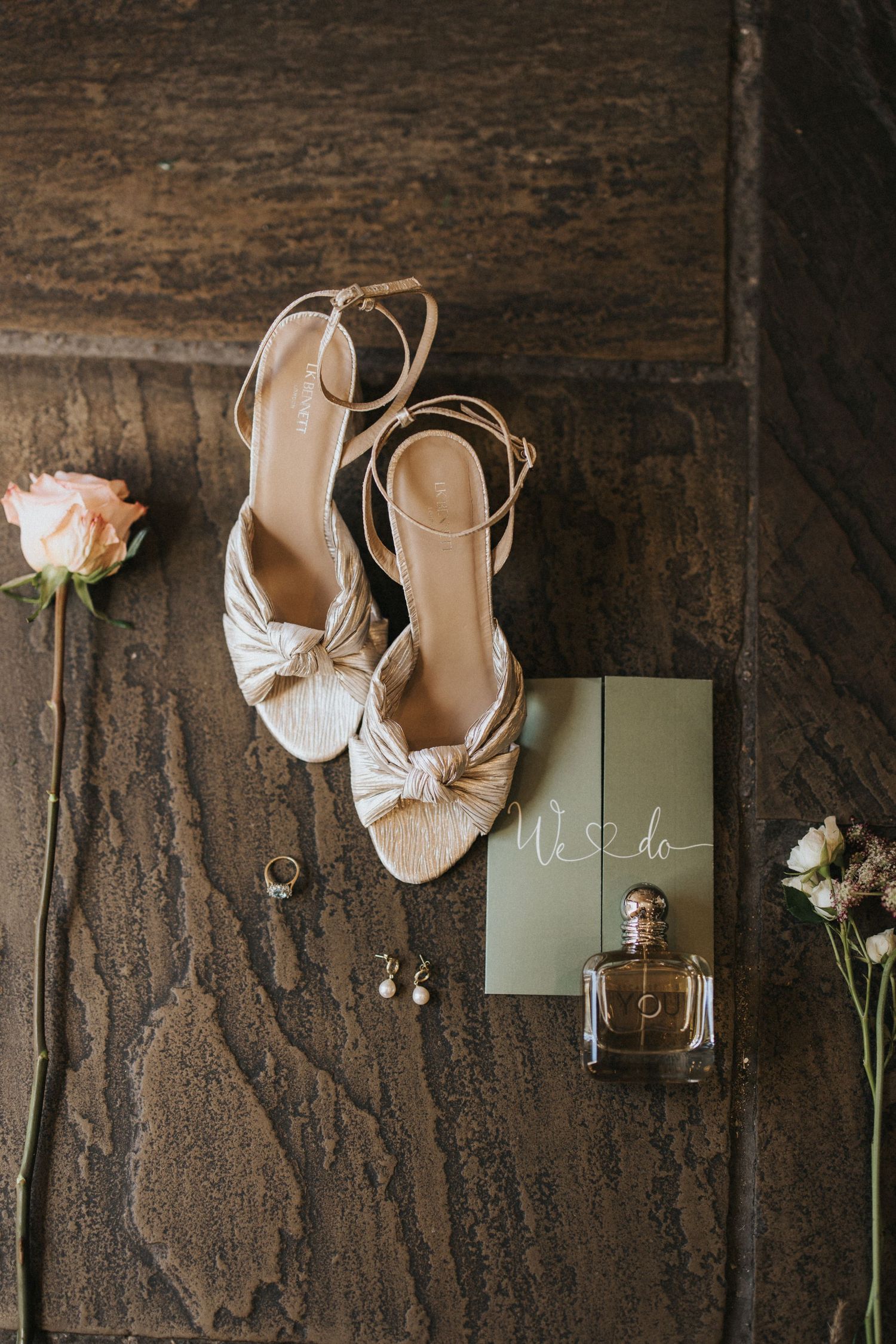 Elegant ivory wedding heels with bow details arranged alongside delicate flowers against dark rustic wood texture.