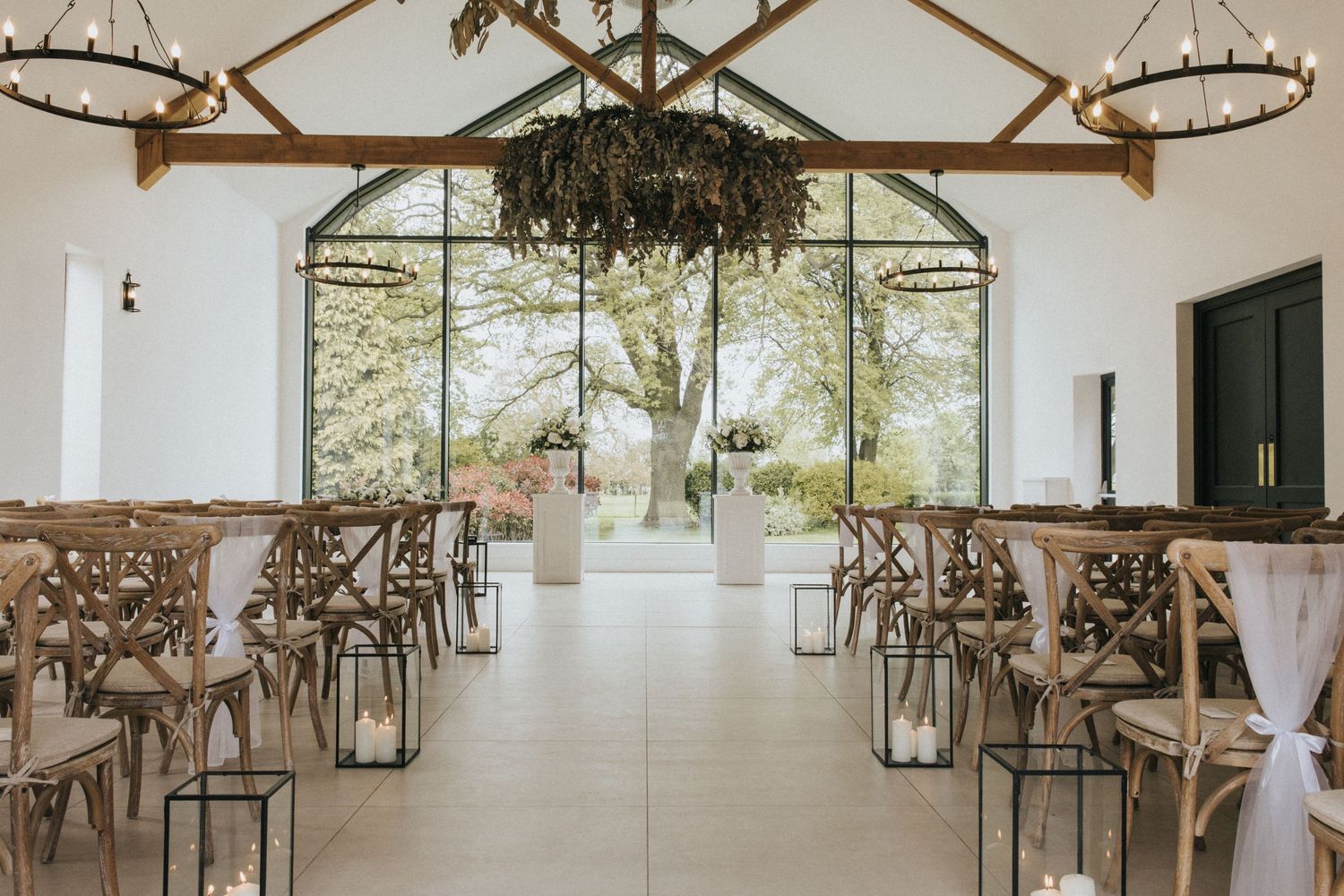 An elegant wedding reception venue with rustic wooden beams, crystal chandeliers, and white-draped decor in a bright space.