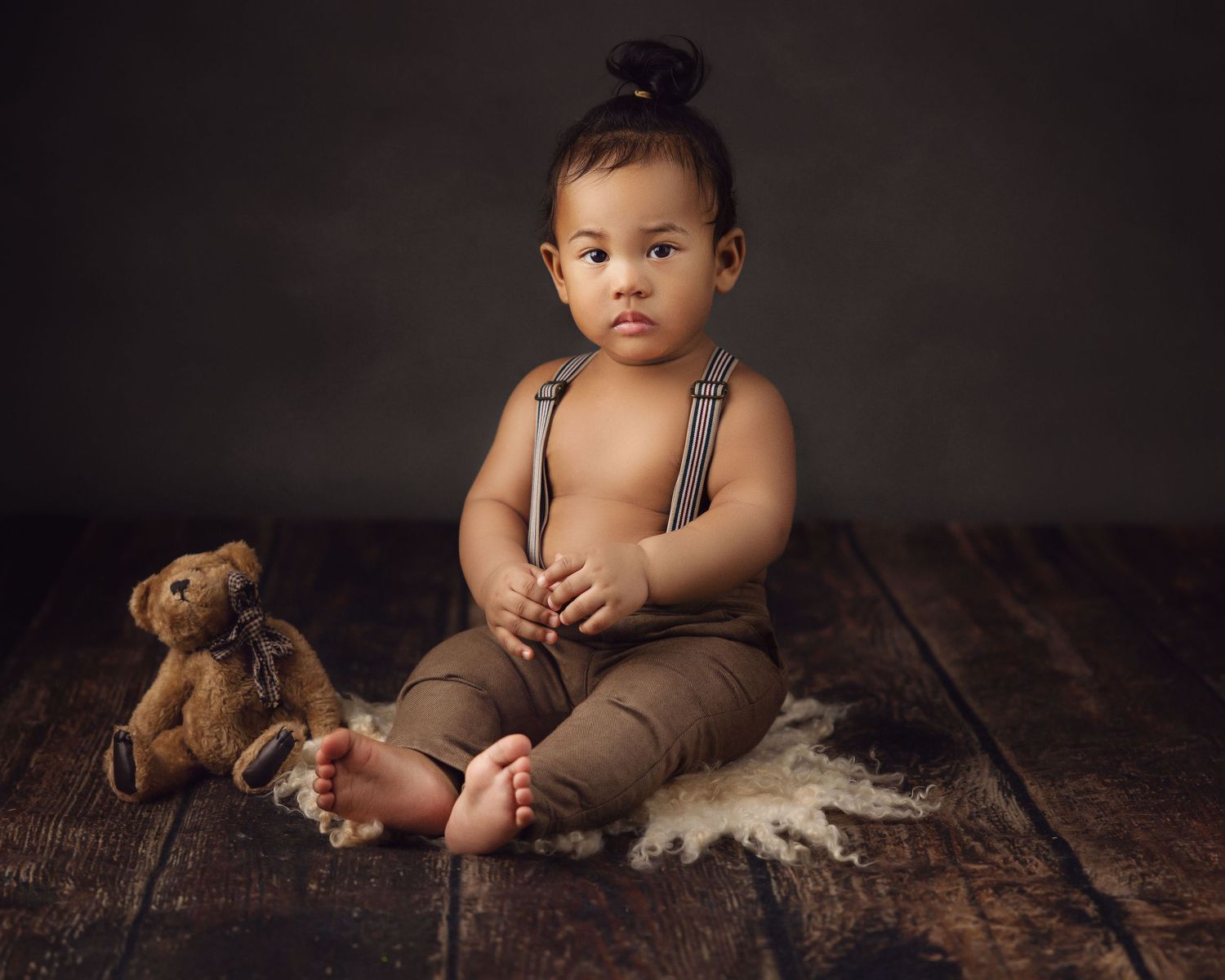Classic portrait of toddler in suspenders with teddy bear on wooden floor.