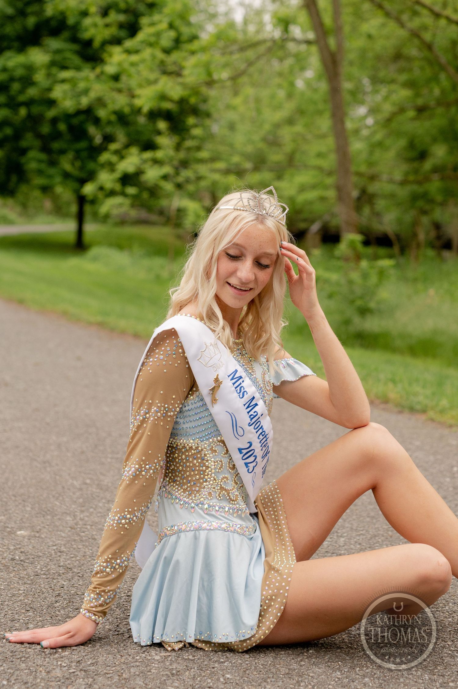 Beauty queen in a detailed white and gold dress poses elegantly on a park pathway.