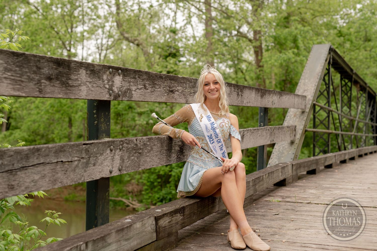 Beauty queen in formal attire poses on a rustic wooden bridge in a park setting.