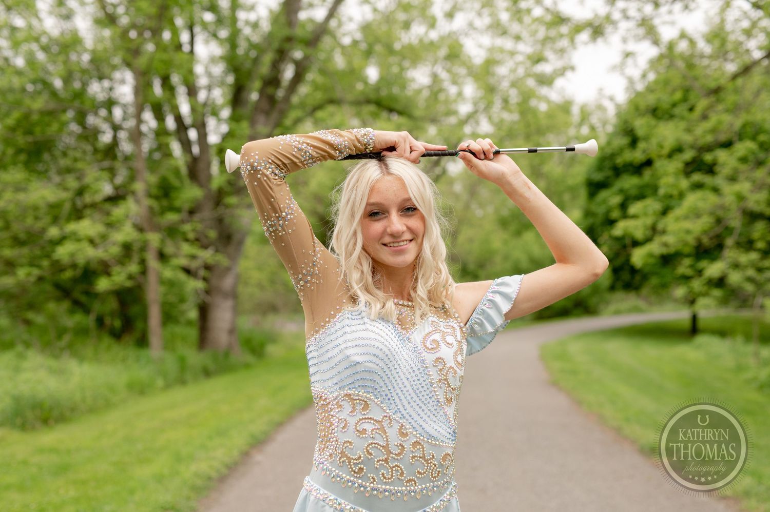 Beauty pageant winner in a sparkly blue and gold dress poses playfully on a path surrounded by green trees.