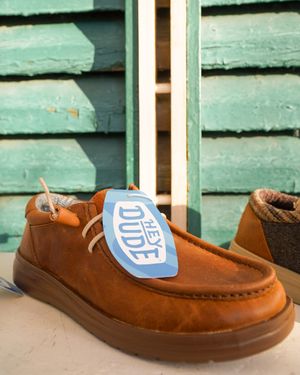 Brown suede slip-on shoes with blue tags displayed against mint green siding.