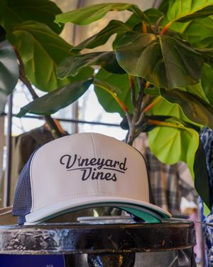 White and navy Vineyard Vines trucker hat displayed beneath green leafy plant.