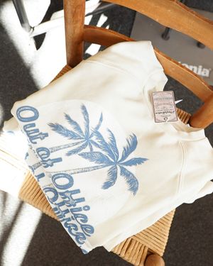 White sweatshirt with blue palm tree design laid on wooden chair.