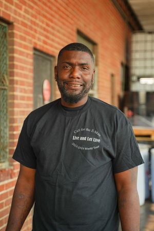A staff member wearing a black t-shirt stands smiling in front of a brick building.