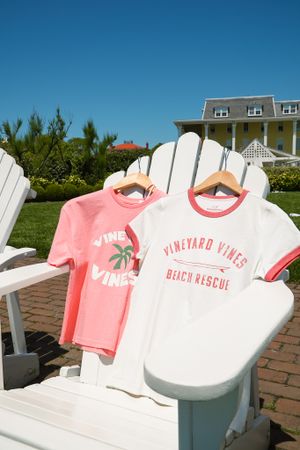 Pink and white beach-themed t-shirts displayed on Adirondack chairs outdoors.
