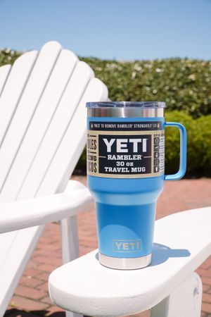 Blue YETI travel mug sitting on white Adirondack chair against sunny backdrop.