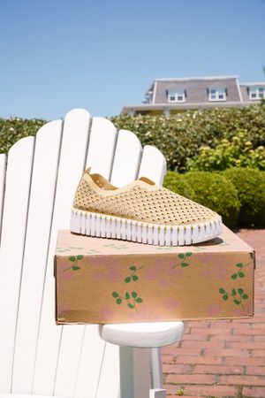 Beige perforated slip-on sneaker displayed on white Adirondack chair with floral box in outdoor setting.