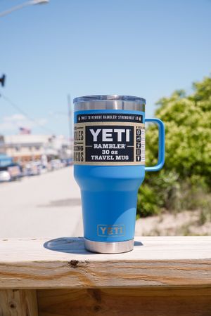 Blue YETI insulated travel mug sitting on wooden railing with ocean view in background.