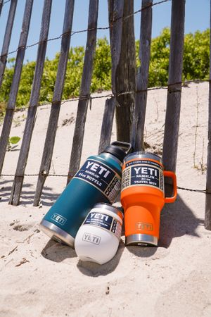 Collection of YETI tumblers in blue, white, and orange arranged on beach sand against wooden fence.