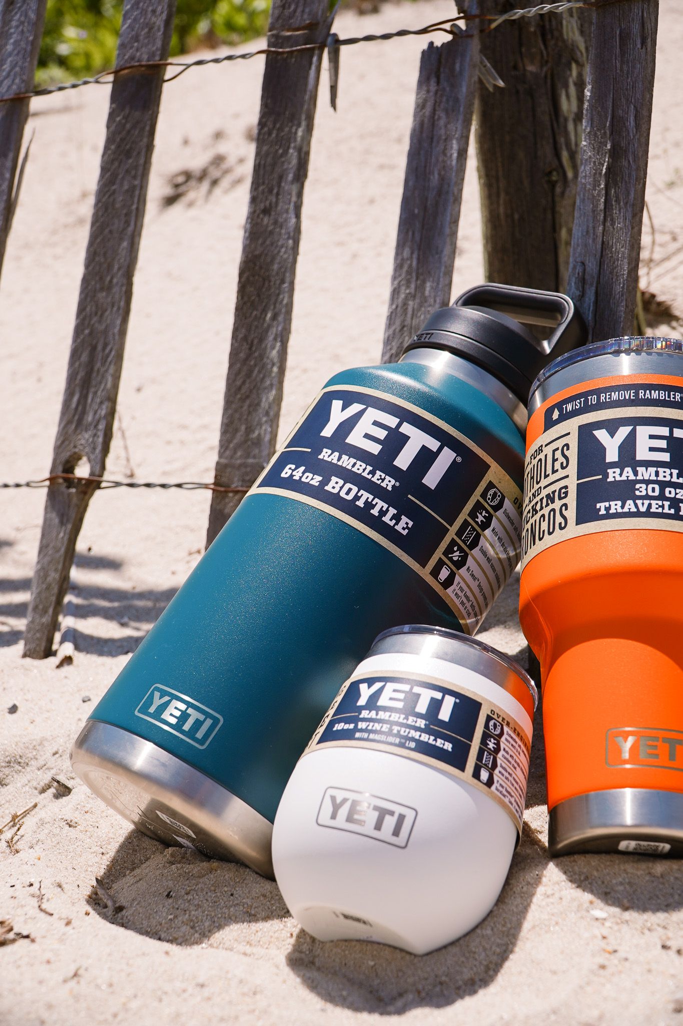 Close-up of YETI drink containers in various sizes and colors positioned against beach fence.