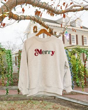 Cream colored sweatshirt with red 'Merry' text hanging on tree branch with holiday lights.
