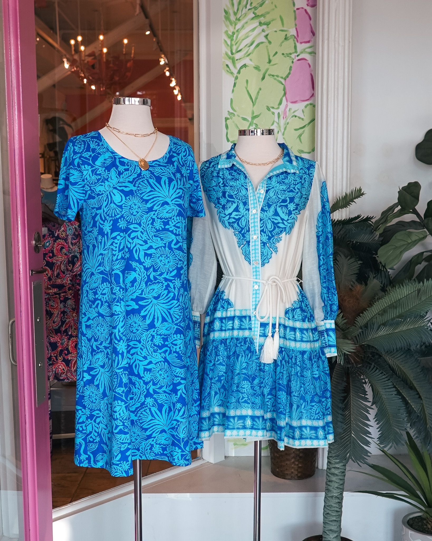 Two blue dresses on display mannequins in a boutique window with tropical wallpaper background.