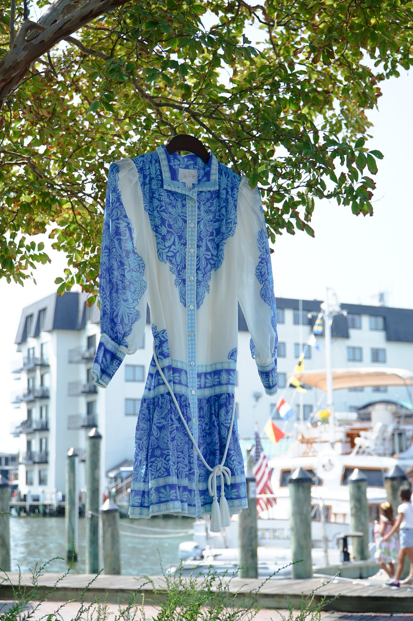 A blue and white patterned dress hanging from a tree branch near a marina with boats in the background.