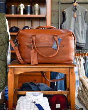 Brown leather duffle bag displayed on wooden shelf with clothing items below.