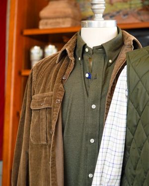 Layered men's clothing display featuring a brown corduroy jacket, olive button-down shirt, and plaid fabric.