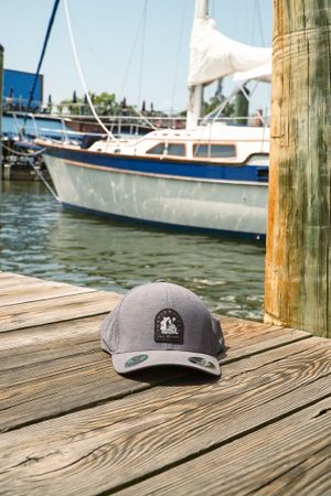 A gray baseball cap with logo sits on wooden dock with sailboat in the background.