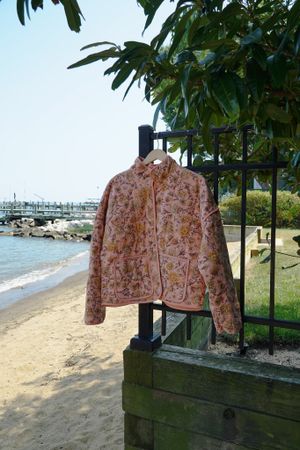 A pink floral jacket hanging on display near a beach with boats visible in the distance.