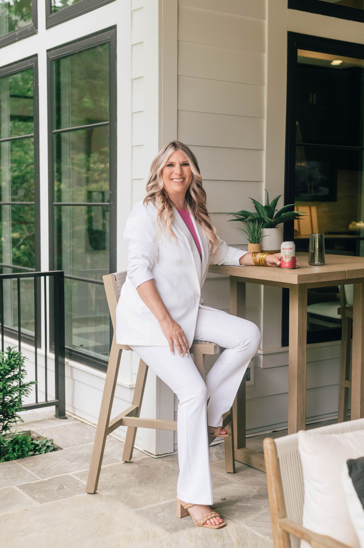 Someone wearing an all-white outfit sits on a wooden stool on an outdoor patio with a small side table and plants. realtor- real estate
