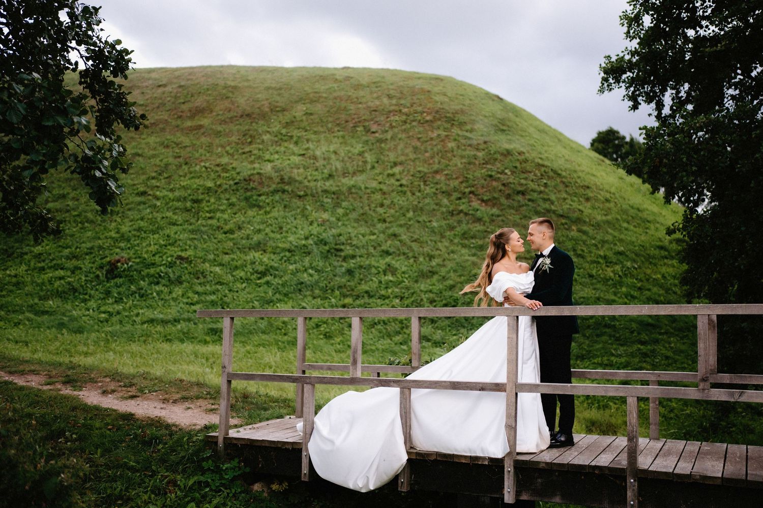 A couple stands on a wooden boardwalk surrounded by trees and grassy hills in romantic wedding photos.
